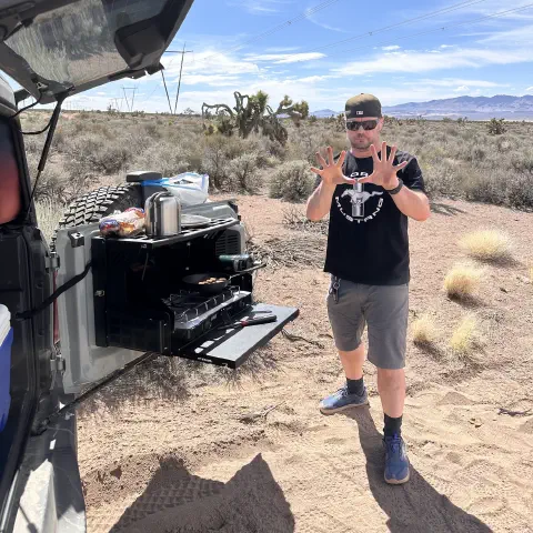 Standing next to open tailgate set up for cooking in the desert with mountains and clear skies in the background.